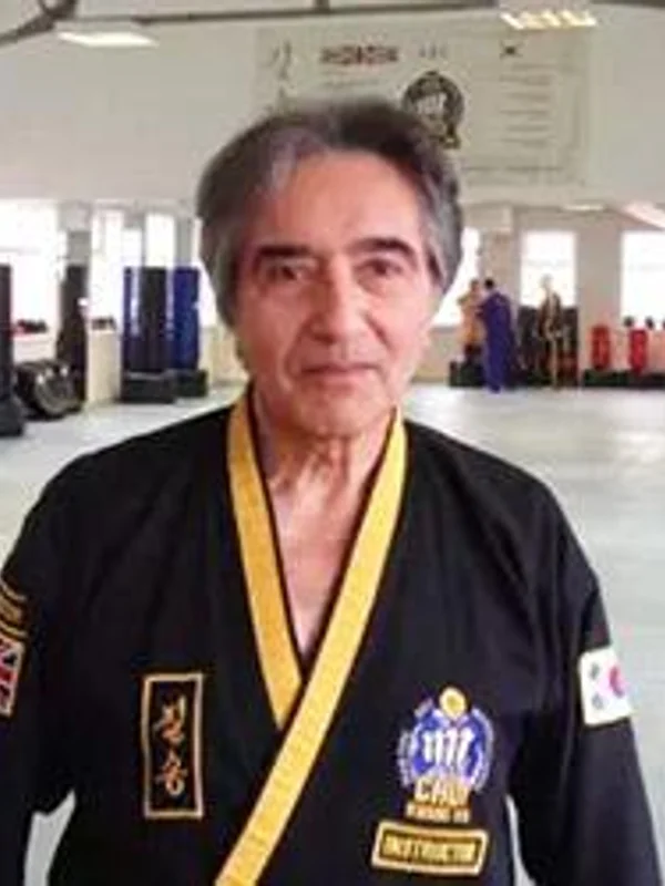 Portrait of an adult instructor wearing a black martial arts uniform with a yellow belt and badge, photographed indoors in what appears to be a training hall wi