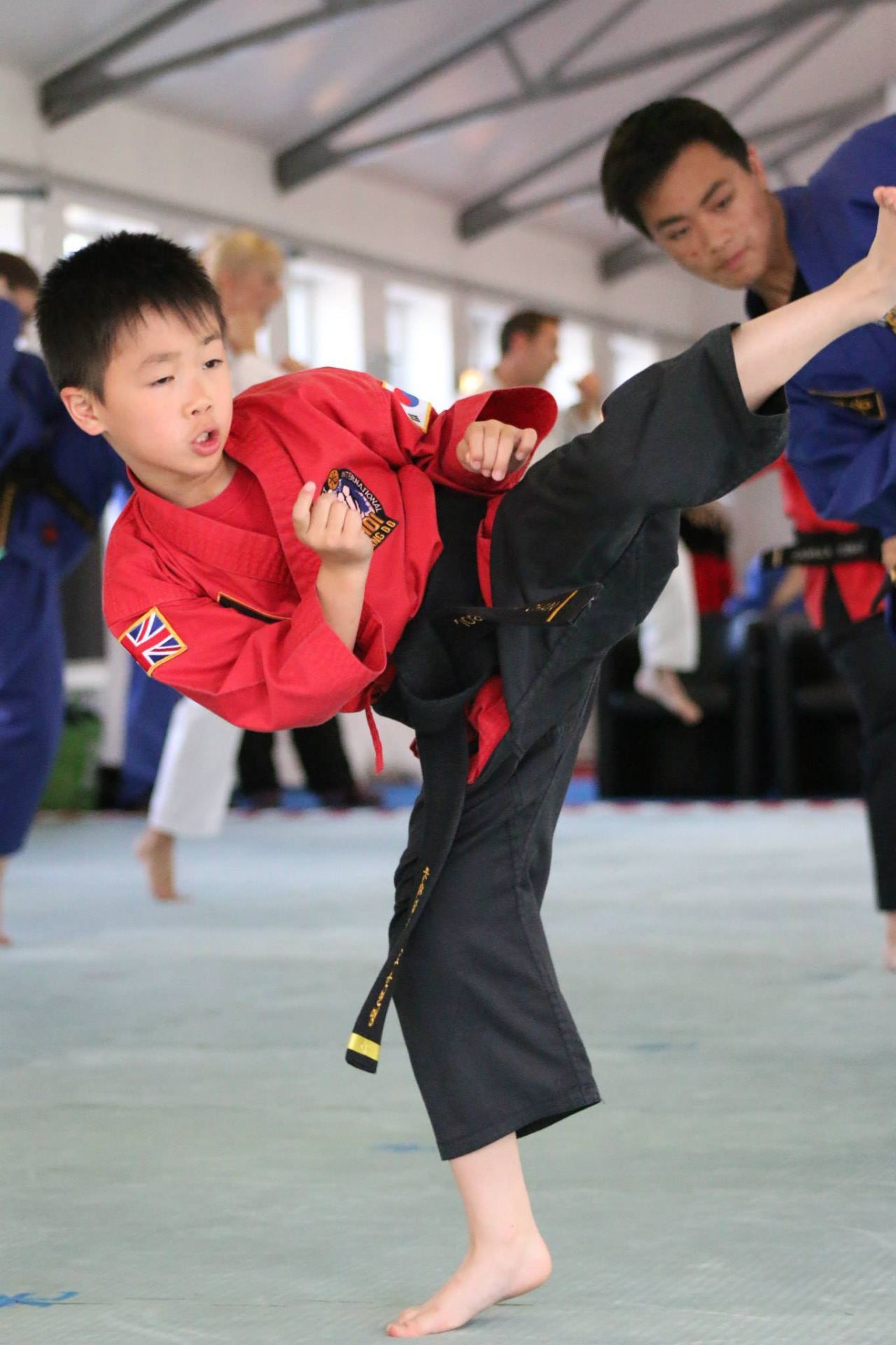 A young child in a red martial arts uniform performs a high kick under the guidance of an instructor in blue in a bright, spacious studio.