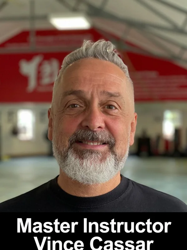 Portrait of an adult man with grey hair and beard, smiling at the camera in what appears to be a large indoor sports or activity hall with red signage and fluor