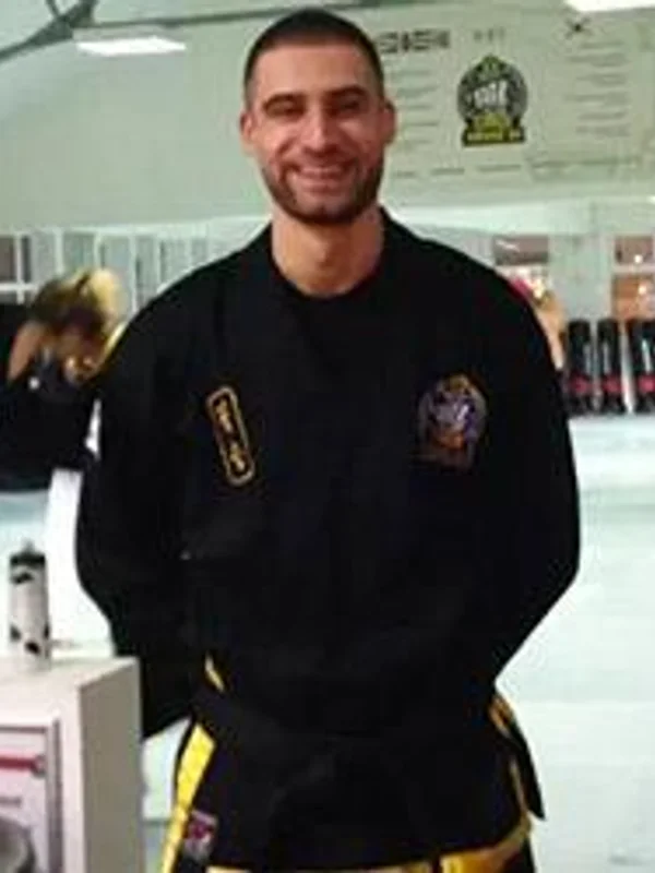 A smiling man in a black martial arts uniform with gold trim stands in what appears to be a studio or gym setting, with posters and other students visible in th