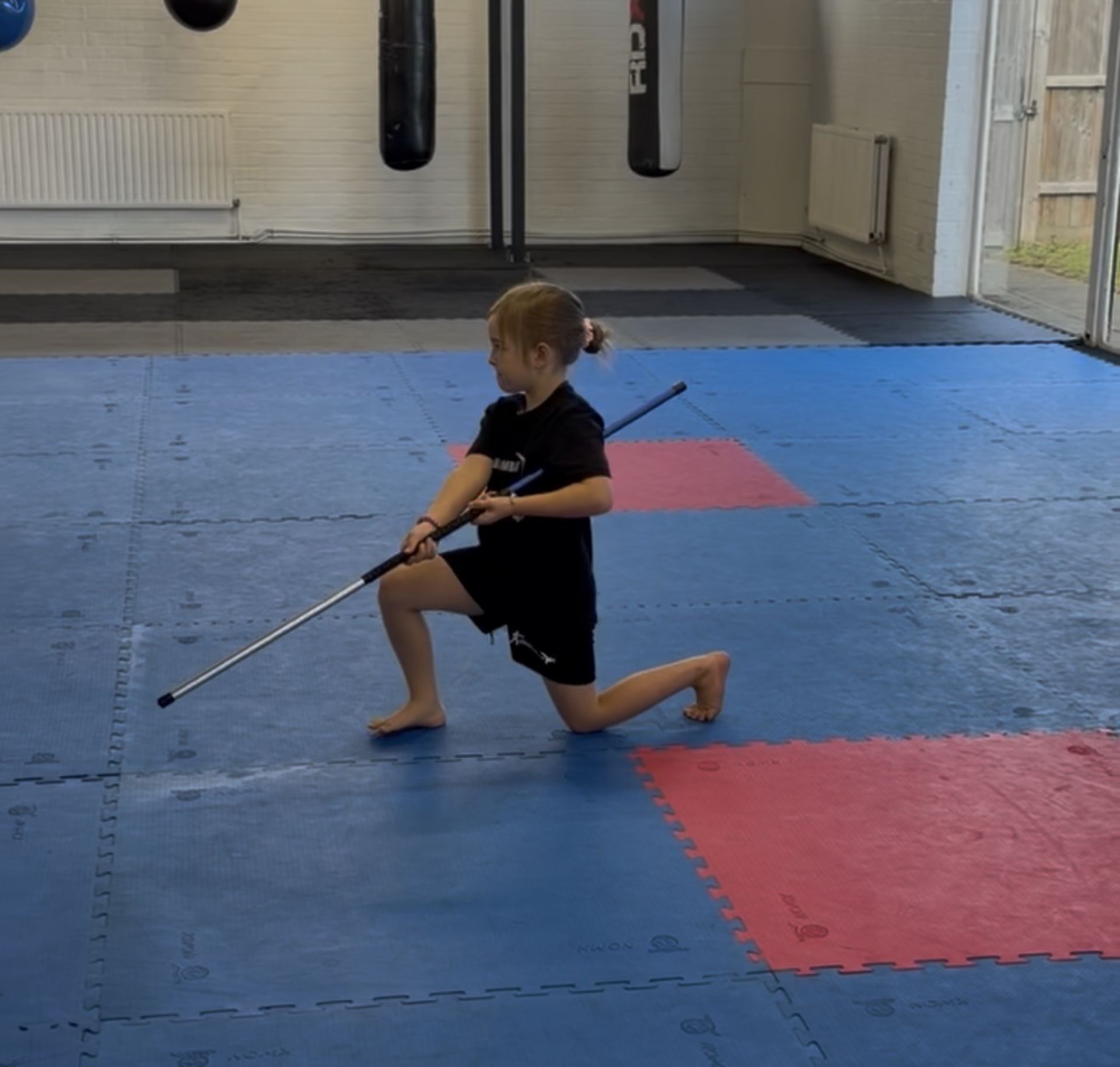 A young child in a black shirt practices with a staff or quarterstaff on a blue padded gym floor with red sections, in a well-lit indoor training space with pun