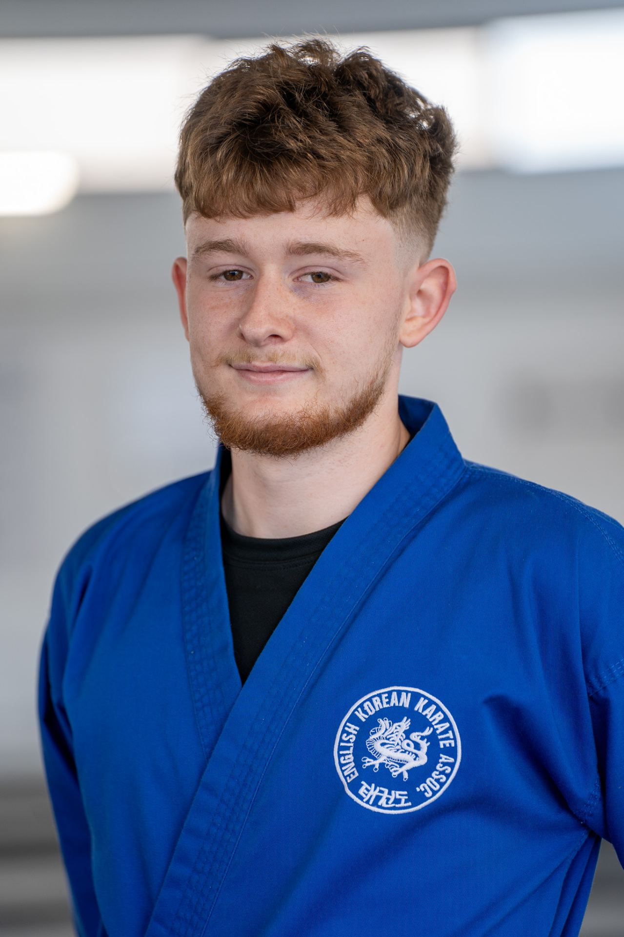 Professional headshot of a young man wearing a blue Korean martial arts uniform (dobok) with an embroidered badge.