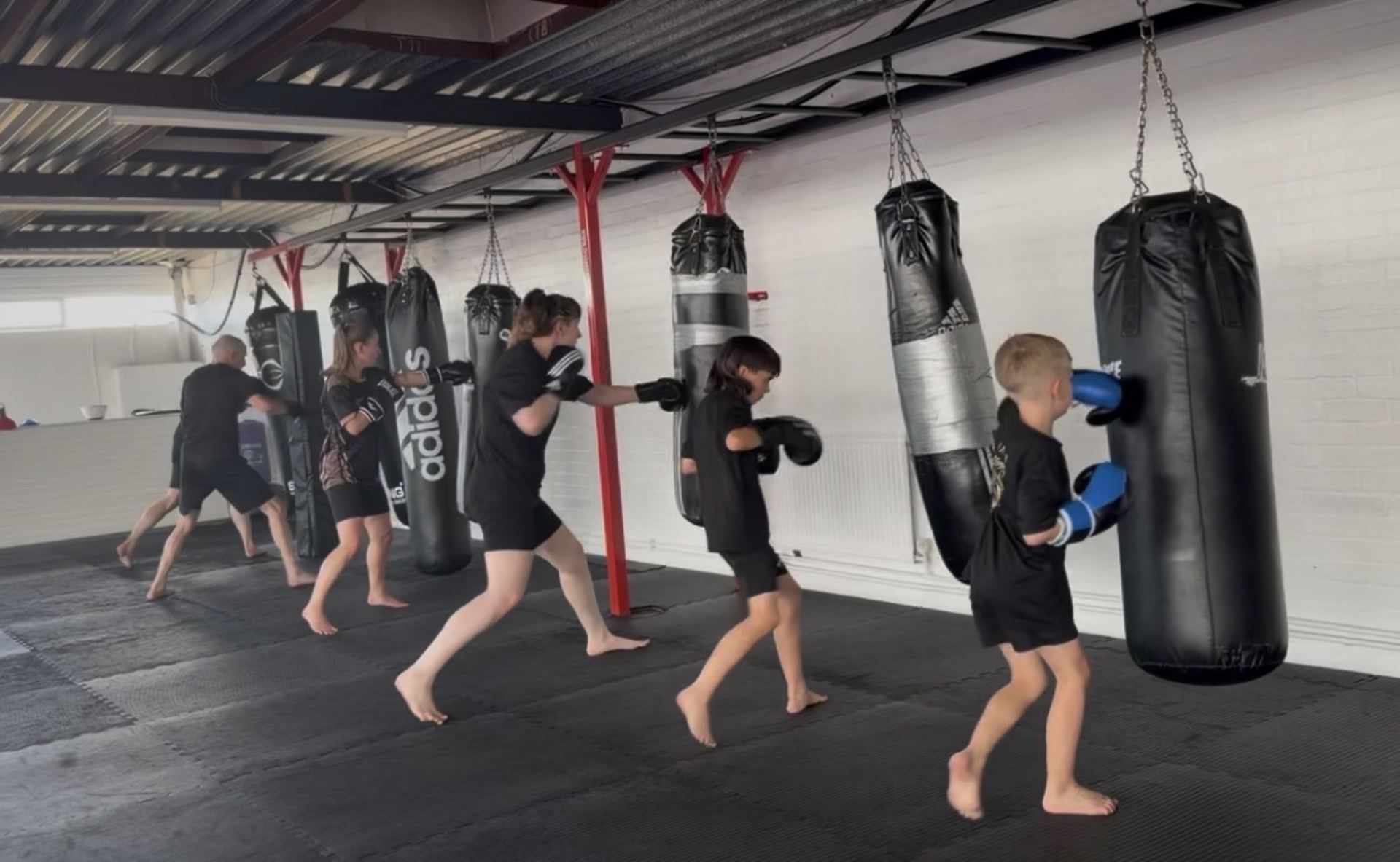 A group of young students in black athletic wear practice boxing with multiple heavy bags suspended from the ceiling in a bright, spacious gym with industrial f