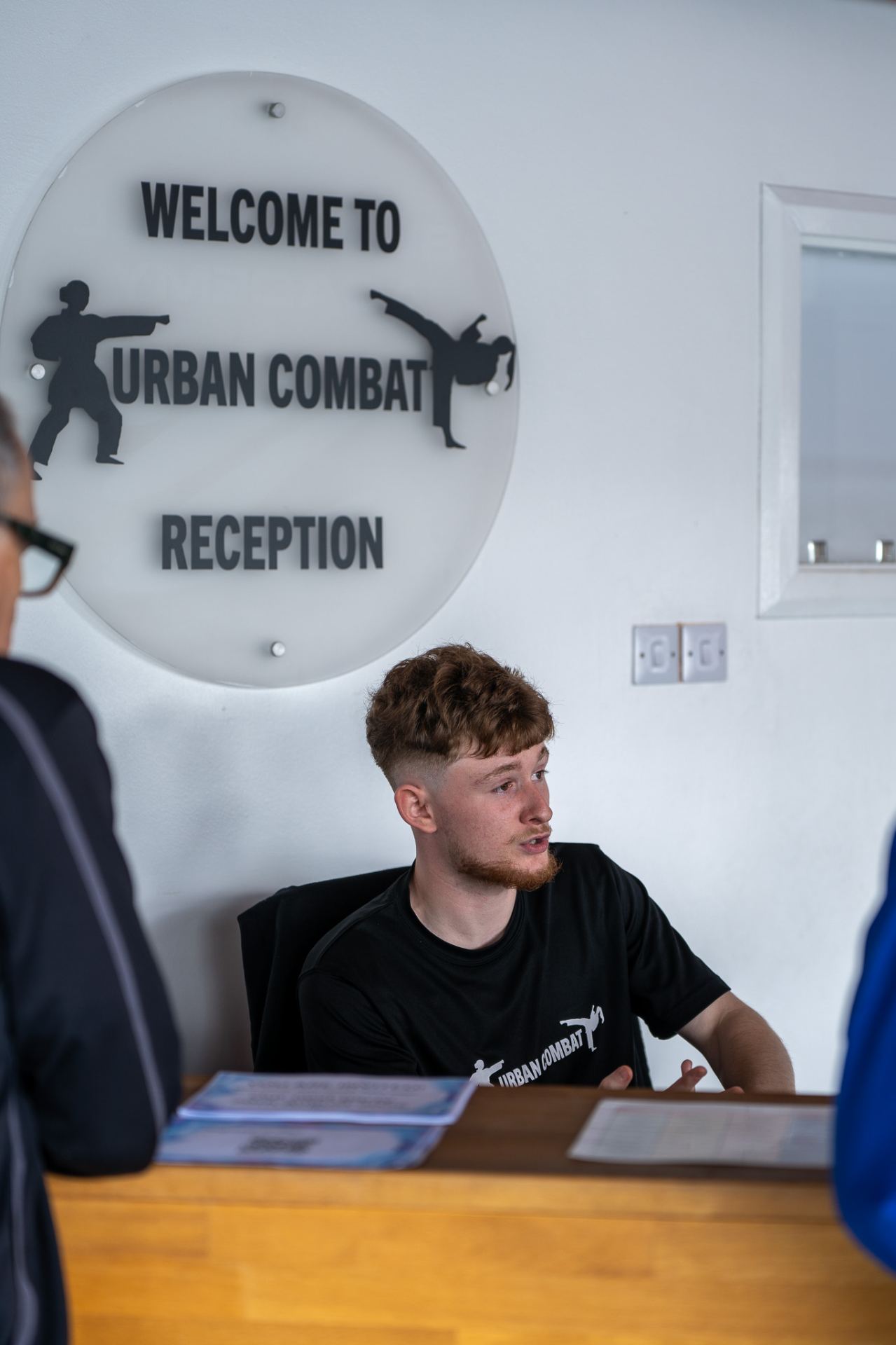 A young man wearing an Urban Combat branded t-shirt sits at a reception desk beneath a large circular sign reading "Welcome to Urban Combat Reception" decorated