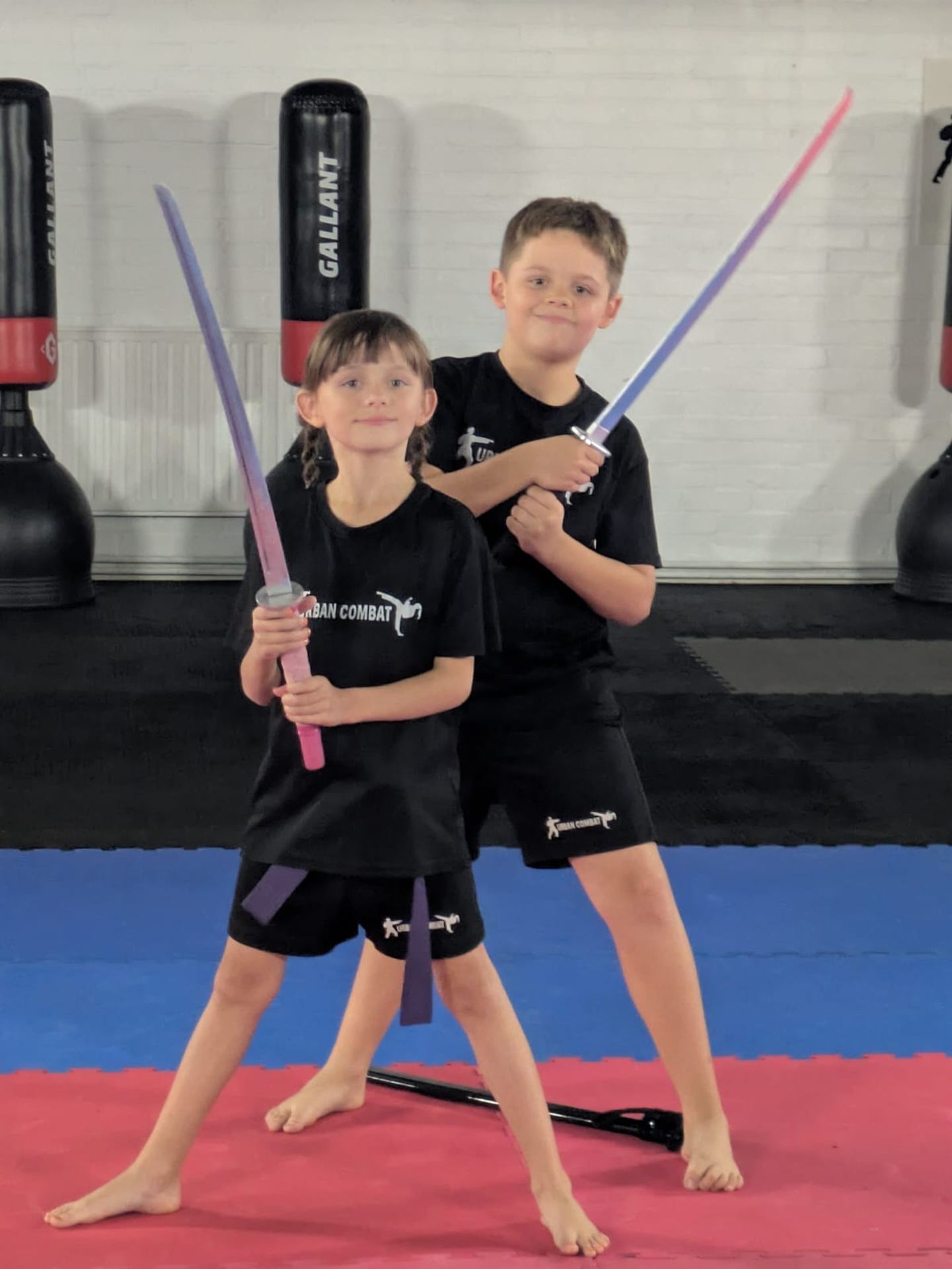 Two young children in black martial arts uniforms holding colorful foam swords on a padded mat in a training studio, with punching bags visible behind them.
