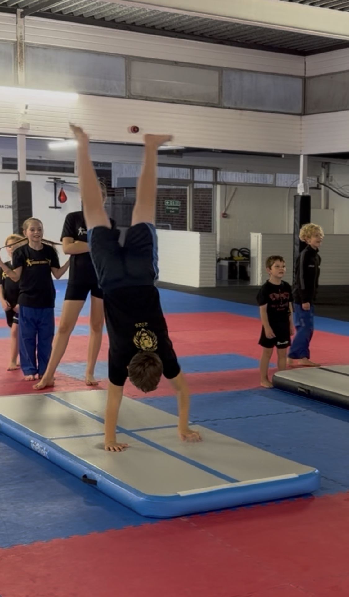 A child performs a handstand on an inflatable air mat in a gym studio, with several other young students watching and waiting their turn.