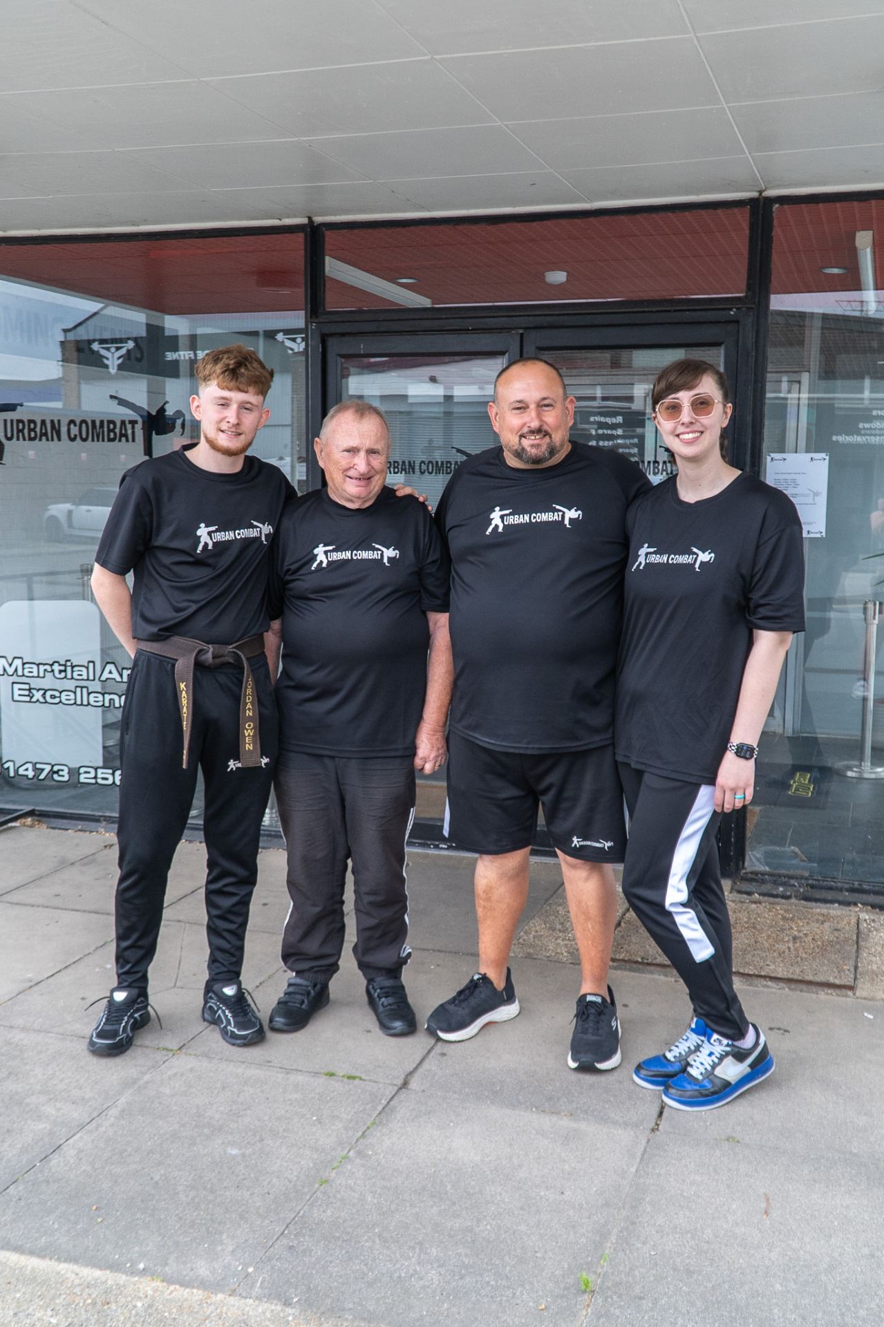 Four people in matching black Urban Combat branded t-shirts stand together outside the school's storefront, smiling at the camera.