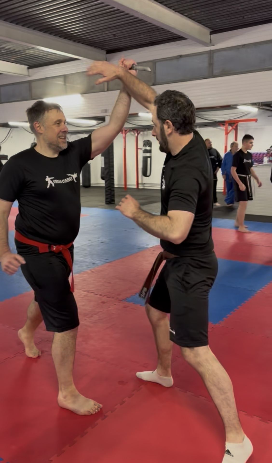 Two adult men in black shirts are engaged in a martial arts or combat sports class on a red and blue padded mat.