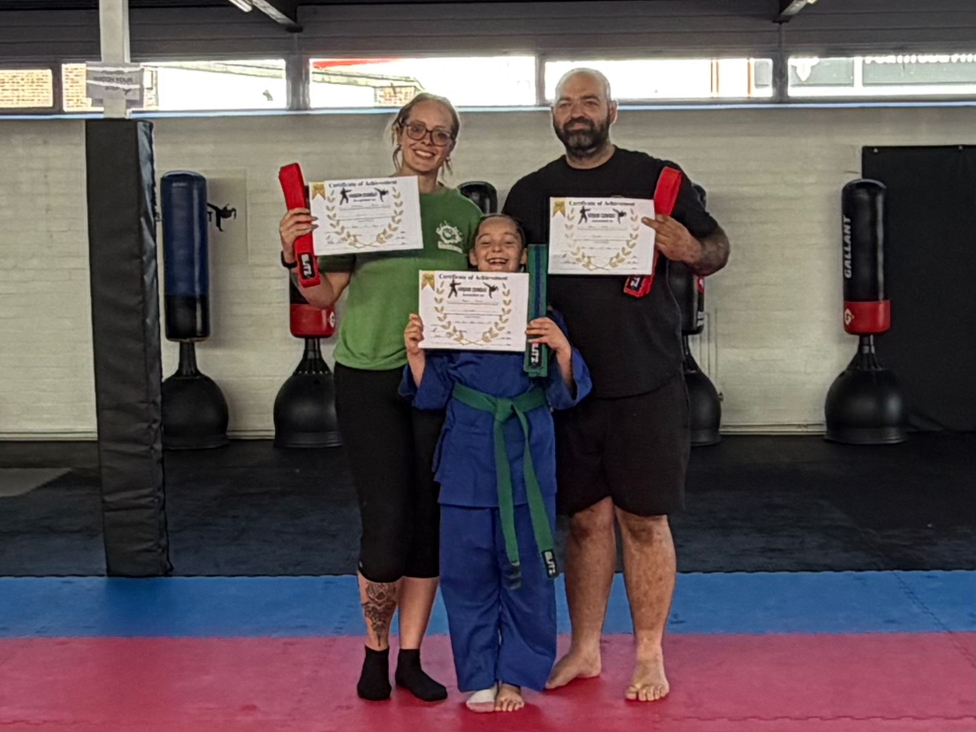 Three people in a martial arts studio display certificates of achievement and red boxing gloves, smiling at the camera.