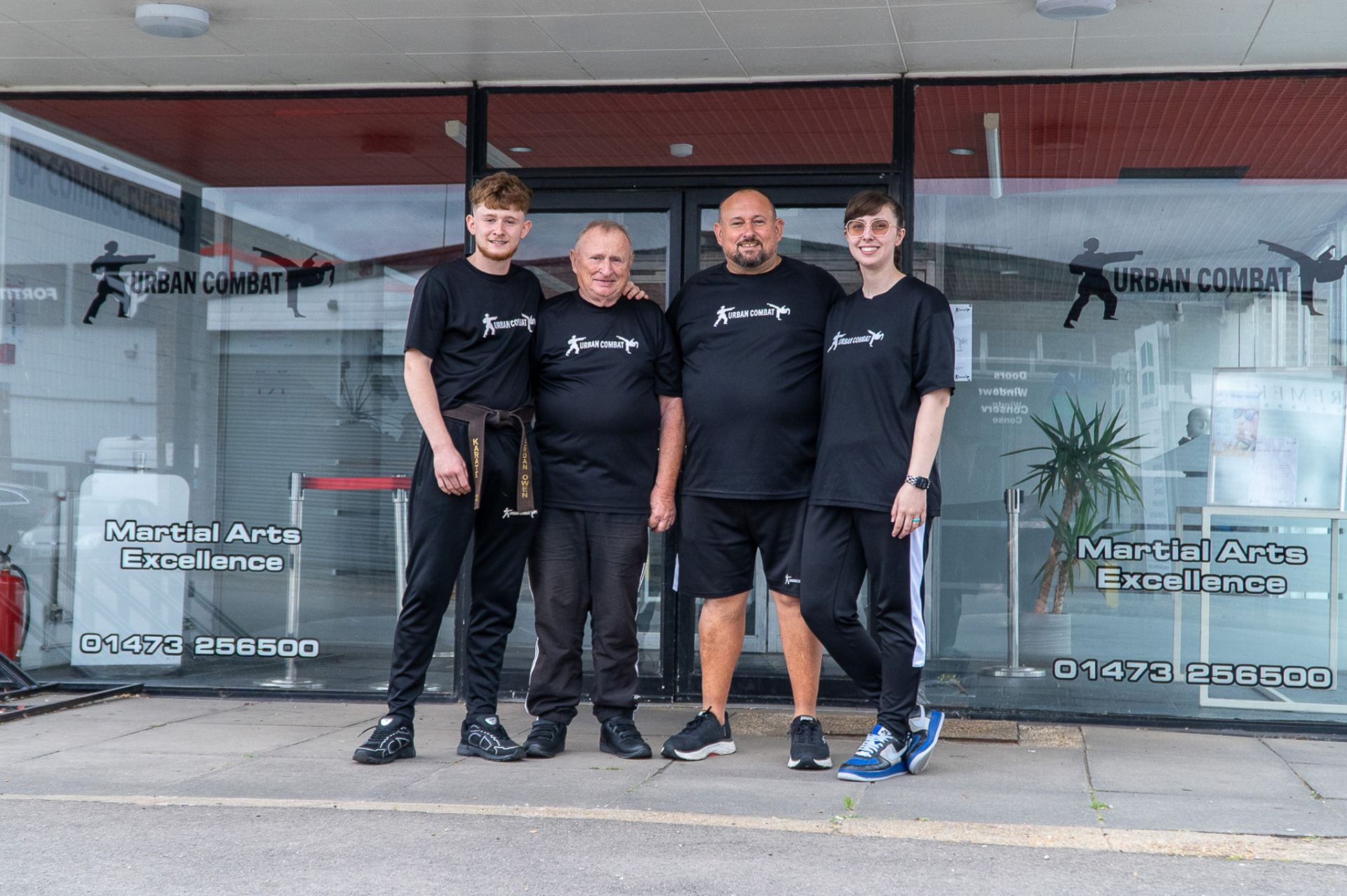 Four people in black branded martial arts uniforms stand together outside a storefront with "Urban Combat" and "Martial Arts Excellence" signage on the windows.