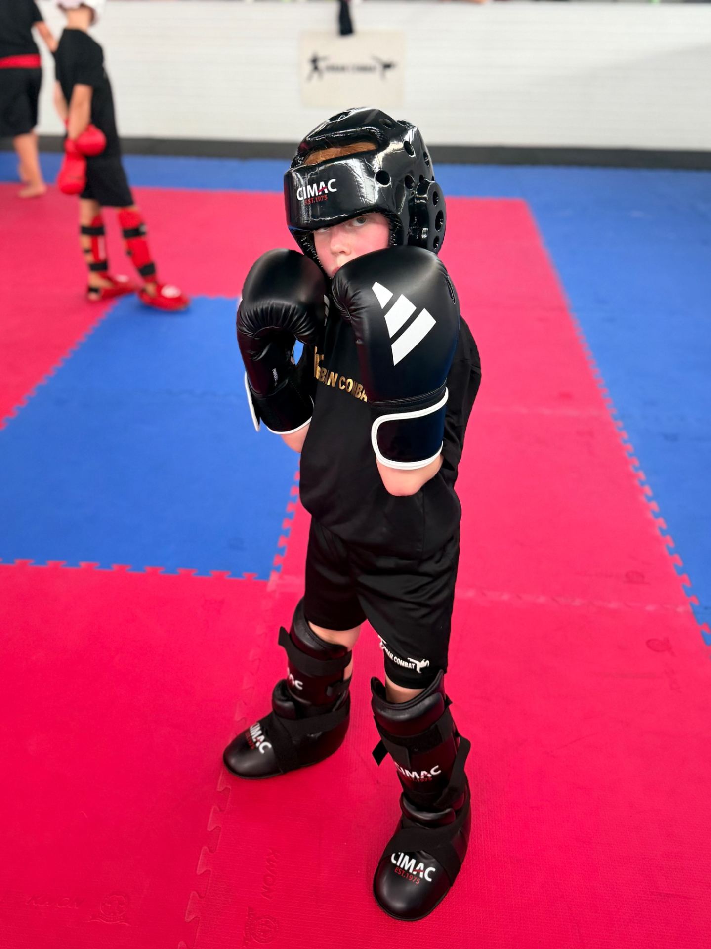 A young child in full protective gear (headguard, gloves, shin guards) stands in a boxing stance on a red and blue padded mat.